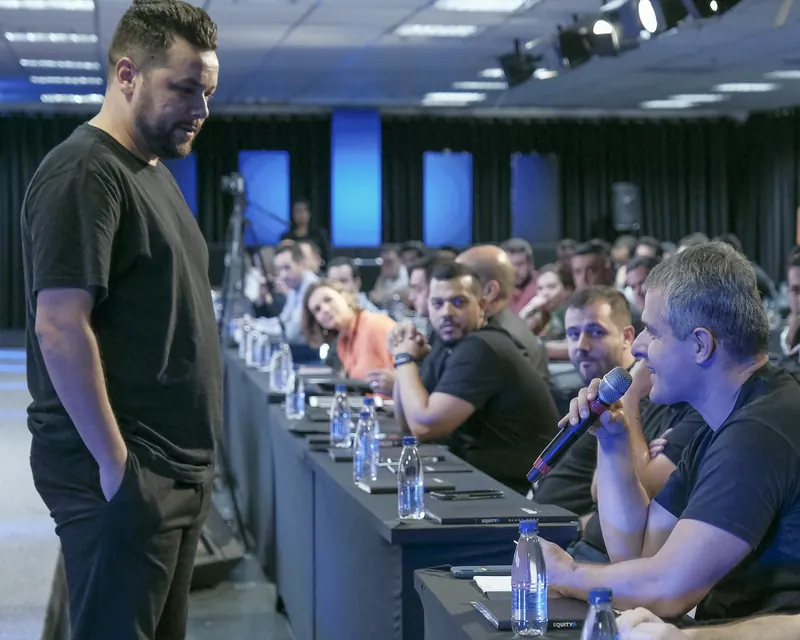 Juvenal de pé, pensativo, ao lado de uma mesa longa de empresários durante um evento