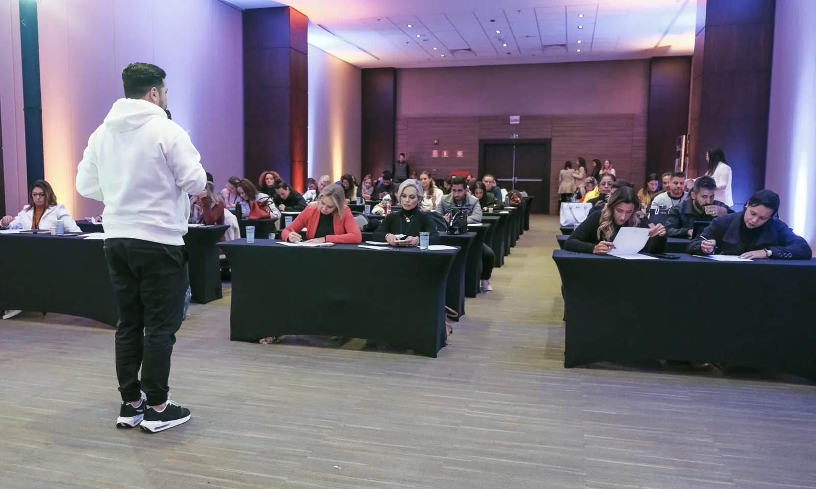 Juvenal de pé, de costas, conduzindo uma sala de empresários sentados em mesas, escrevendo